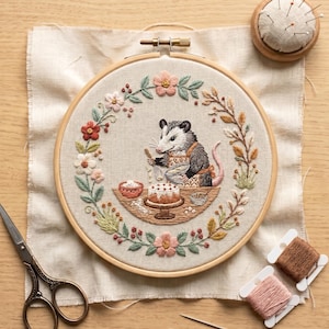 May include: An embroidery hoop containing a fabric piece with a stitched opossum wearing an apron and baking a cake, surrounded by a floral wreath. The scene includes a pair of scissors, two spools of thread, two needles, and a pincushion nearby.