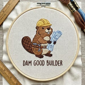 Op de afbeelding: Geborduurde hoepelkunst met een cartoon bever met een gele helm en gereedschapsriem, die bouwtekeningen vasthoudt. De tekst "DAM GOOD BUILDER" is eronder geborduurd. Bruin en blauw borduurgaren en een houten liniaal zijn zichtbaar.