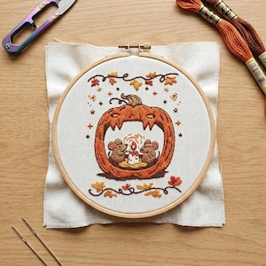 May include: An embroidery hoop containing a fabric piece with a stitched design of a carved orange pumpkin. Inside the pumpkin, two brown mice sit beside a lit candle. The design is surrounded by small orange and brown autumn leaves and stars.