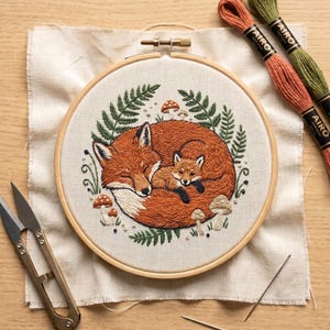 May include: An embroidery hoop containing a fabric piece with a stitched design of a large orange fox curled around a small fox kit, surrounded by green ferns and small mushrooms. Includes two skeins of Airo embroidery floss and metal scissors.