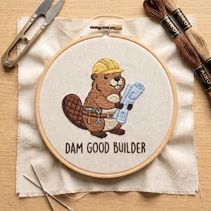May include: An embroidery hoop containing a beige fabric with a stitched brown beaver wearing a yellow hard hat and a tool belt, holding blue blueprints. Below the beaver, the text DAM GOOD BUILDER is stitched in brown thread. Metal scissors and two brown embroidery floss skeins are present.