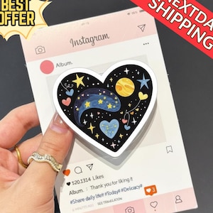 May include: A heart-shaped phone grip featuring a celestial design with stars, a moon, and a galaxy pattern. The grip is held over a mock Instagram post. The image also includes the text "BEST OFFER" and "NEXTDAY SHIPPING".