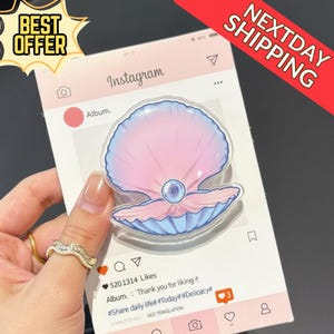 May include: A clear, seashell-shaped phone grip with pink and blue hues, and a pearl in the center. The grip is held in front of a pink Instagram-themed card. The card has the text "Album" and "5201314 Likes".