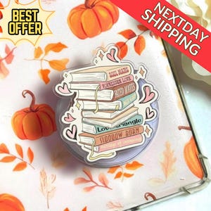 May include: A clear phone grip featuring a stack of books with titles like "Soul Mates" and "Forbidden Love." Pink hearts and sparkles surround the books. The background includes pumpkins and fall foliage.