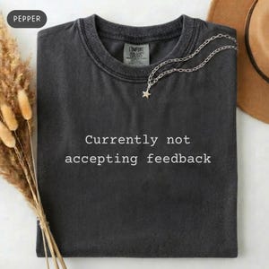 May include: A dark gray t-shirt with the text "Currently not accepting feedback" in white. A silver necklace with a star pendant rests on the shirt. The shirt is folded and displayed on a neutral background.