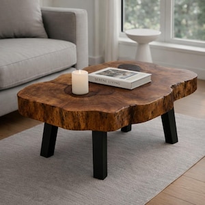May include: A live-edge coffee table with a natural wood grain finish. The table has black metal legs and a thick, organic shape. A white candle and a book titled "SERENE LIVING" rest on the table.