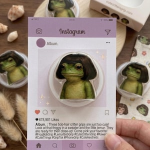 May include: A clear phone grip featuring a green frog with a black bob haircut, set against a light purple Instagram-themed background. The text on the card reads "Album." and "Instagram."
