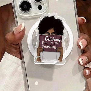 Könnte beinhalten: Transparente Handyhülle mit einem weißen Pop-Socket, das ein Cartoon-Bild einer Person mit Afro-Frisur zeigt, die ein Buch hält. Das Buch trägt die Aufschrift "Go away I'm reading". Die Handyhülle wird von einer Person mit manikürten Nägeln gehalten.