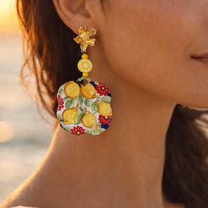 Può includere: Orecchini a bottone dorati con motivo floreale e pendente. Il pendente presenta una perlina a fetta di limone, un ciondolo a forma di fiore e una grande goccia fantasia con limoni e fiori rossi. Un accessorio vivace.