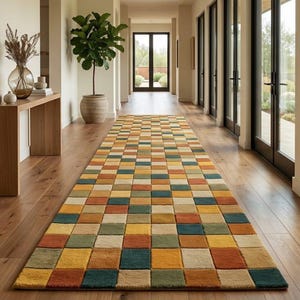 Puede incluir: Una alfombra rectangular con un patrón geométrico de cuadrados en tonos naranja, amarillo, verde y azul. La alfombra está colocada en un pasillo con suelo de madera y grandes ventanales.
