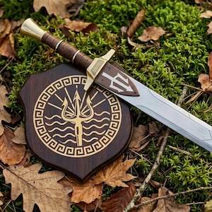 May include: A decorative sword and shield set. The sword has a silver blade, a brown handle, and a gold-coloured hilt. The shield is dark brown with a gold trident design. The set is displayed on a bed of green moss and fallen leaves.