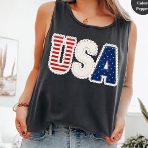 May include: A dark gray sleeveless tank top featuring the letters USA in a decorative font. The U has red and white horizontal stripes, the S is solid white, and the A is dark blue with white polka dots. Each letter has a white scalloped trim.