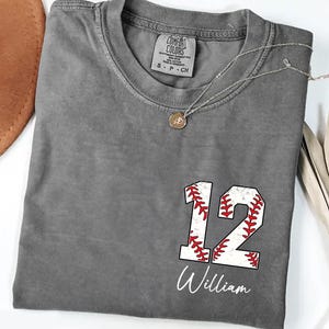 May include: A gray Comfort Colors t-shirt with the number 12 in a baseball design and the name "William" in white script. A silver necklace with a pendant is draped across the shirt.