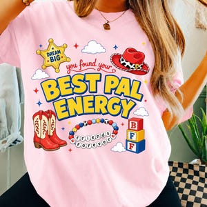 May include: A light pink t-shirt featuring a graphic print with the text "you found your BEST PAL ENERGY". The design includes a yellow sheriff star, a red cowboy hat, red cowboy boots, a beaded bracelet with "friends forever", and blocks.