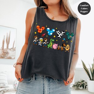 May include: A dark gray sleeveless tank top featuring a graphic print of cartoon characters and Mickey Mouse-shaped balloons. The design includes a cowboy, an astronaut, a cowgirl, a green dinosaur, a pink pig, a horse, and a potato character.