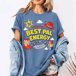 May include: A blue t-shirt featuring a graphic print with the text "you found your BEST PAL ENERGY". The design includes a yellow sheriff star, a red cowboy hat, red cowboy boots, a beaded bracelet with "friends forever" text, and blocks.