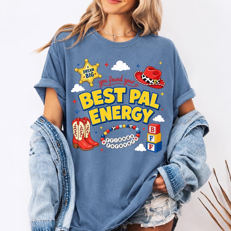 May include: A blue t-shirt featuring a graphic print with the text "you found your BEST PAL ENERGY". The design includes a yellow sheriff star, a red cowboy hat, red cowboy boots, a beaded bracelet with "friends forever" text, and blocks.