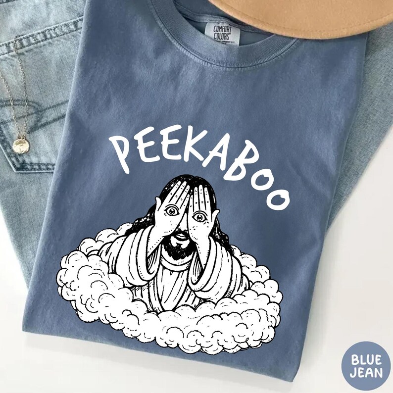 May include: Blue jean-colored t-shirt with the word "PEEKABOO" in white script above a black and white illustration of a figure with hands covering their eyes. The figure is surrounded by a cloud-like design.