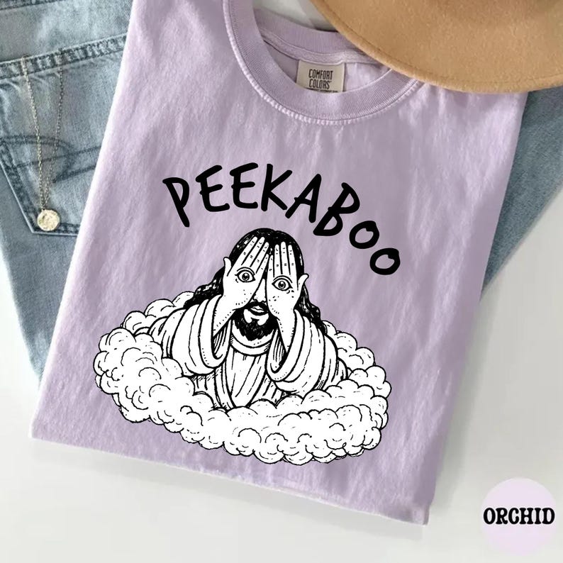 May include: Orchid-colored t-shirt with the word "PEEKABOO" in black above a black and white illustration of a figure peeking through their fingers. The shirt is made of a soft material.
