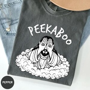 May include: Dark gray t-shirt with the word "PEEKABOO" above a white illustration of a figure with hands covering their eyes. The figure is surrounded by a cloud-like design. The shirt is folded on top of blue denim.