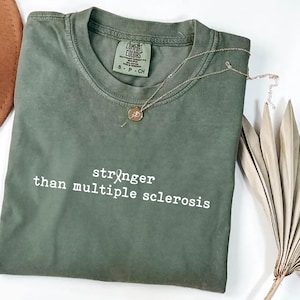 May include: An olive green t-shirt with the text "stronger than multiple sclerosis" in white. A gold necklace with a pendant is draped across the shirt. The shirt has a Comfort Colors tag.