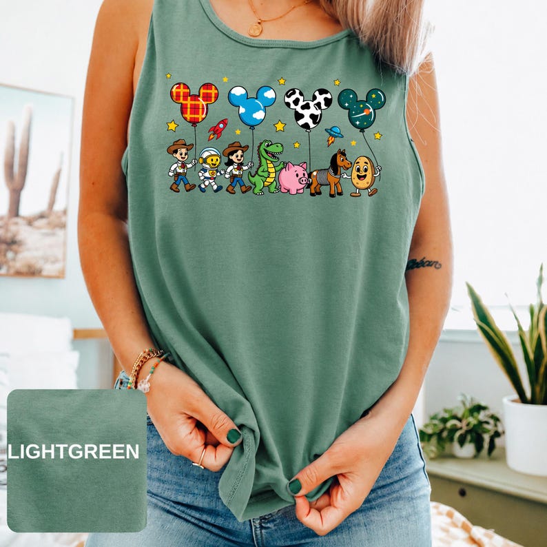 May include: A sage green sleeveless tank top featuring a graphic print of cartoon characters including a cowboy, astronaut, cowgirl, green dinosaur, pink pig, horse, and potato. Above them are four Mickey Mouse-shaped balloons with various patterns.