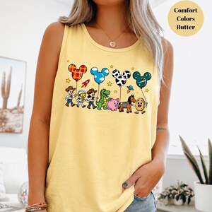 May include: A butter yellow tank top featuring a graphic print of cartoon characters including a cowboy, astronaut, cowgirl, green dinosaur, pink pig, horse, and potato. Above them are four Mickey Mouse-shaped balloons with plaid, cloud, cow, and space patterns.