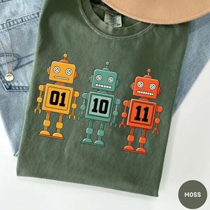 May include: A moss green t-shirt featuring three cartoon robots in yellow, teal, and orange. Each robot has a number on its chest: 01, 10, and 11. The shirt is made of a soft material and is suitable for casual wear.