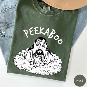 May include: A moss green t-shirt with the word "PEEKABOO" in white script above a black and white illustration of a figure with hands covering their eyes. The figure is surrounded by a cloud-like design. The shirt is displayed on a light blue denim surface.