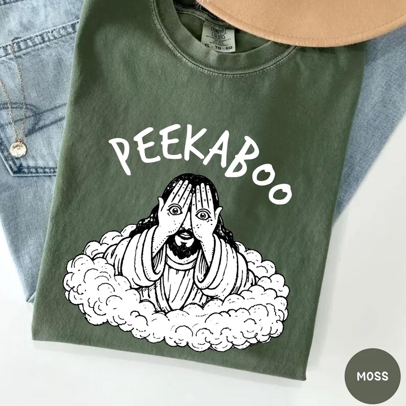 May include: A moss green t-shirt with the word "PEEKABOO" in white script above a black and white illustration of a figure with hands covering their eyes. The figure is surrounded by a cloud-like design. The shirt is displayed on a light blue denim surface.