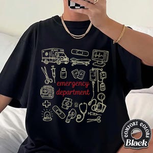 May include: Black t-shirt with a cream-colored graphic of medical equipment and the words "emergency department." The design includes an ambulance, heart monitor, stethoscope, and other medical symbols. The shirt has a crew neck and short sleeves.