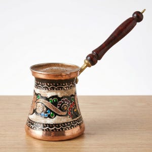 May include: A copper coffee pot with a dark wooden handle and ornate floral designs in red, green, blue, and gold. The pot is filled with coffee and sits on a wooden surface.