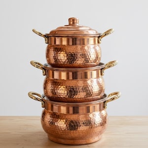 May include: A stack of three copper pots with hammered textures and brass handles. The top pot has a lid with a small knob. The pots are arranged on a wooden surface, with a neutral background.