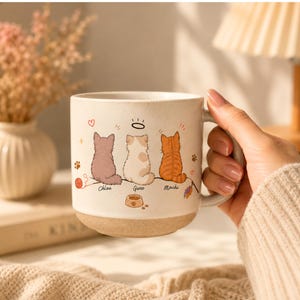 May include: A ceramic mug with a speckled cream finish featuring a graphic of three cats sitting side-by-side. The cats are labeled Chloe, Gwen, and Mochi. The design includes small hearts, stars, paw prints, a ball of yarn, and a food bowl.