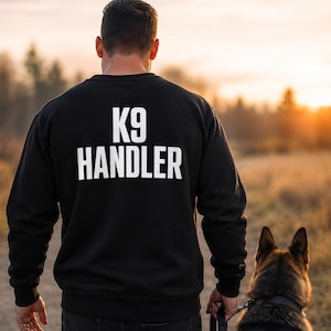 May include: A black long-sleeve sweatshirt featuring the white text K9 HANDLER printed in a bold, sans-serif font across the upper back. The garment has a crew neckline, long sleeves with ribbed cuffs, and a ribbed hemline at the waist.