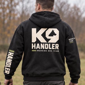 May include: A black hooded sweatshirt featuring a large beige K9 HANDLER graphic on the back, with the text WORKING DOG TEAM below it. The right sleeve displays the word HANDLER in beige, and the left sleeve has WORKING DOG TEAM text.
