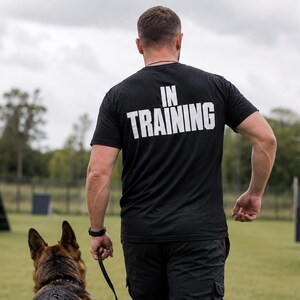以下が含まれることがあります： 背面に白い文字でIN TRAININGとプリントされた黒い半袖Tシャツ。黒いカーゴパンツと黒いブーツを着用した人物が、黒いリードをつけたジャーマンシェパードと一緒にフィールドを歩いている様子。