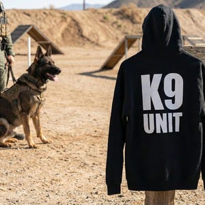 May include: A black hoodie with the white text "K9 UNIT" on the back. The hoodie is displayed outdoors. In the background, a police dog and handler are training on an agility course, with a police car visible.