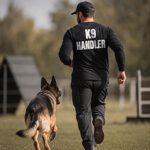 Può includere: Una maglia nera a maniche lunghe con la scritta bianca K9 HANDLER stampata sul retro, abbinata a pantaloni cargo neri e un berretto da baseball nero. La maglia presenta un girocollo e maniche lunghe, indossata da una persona che corre accanto a un pastore tedesco.