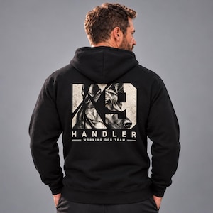 May include: A black hooded sweatshirt featuring a large graphic on the back. The design includes the text K9, HANDLER, and WORKING DOG TEAM, along with a detailed illustration of a German Shepherd dog's face integrated into the lettering.