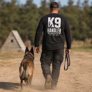 May include: A black long-sleeve shirt featuring white text on the back that reads K9 HANDLER WORKING DOG TEAM TRAIN TRACK CONTROL with a graphic of a person and a dog. The shirt is worn with black cargo pants and a black baseball cap.