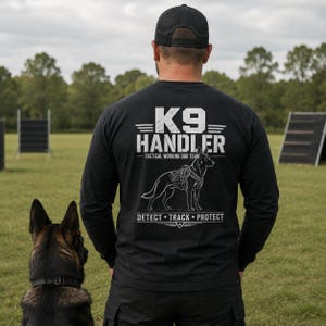 May include: A black long-sleeve shirt featuring a white graphic on the back with the text K9 HANDLER TACTICAL WORKING DOG TEAM and DETECT TRACK PROTECT, accompanied by an illustration of a dog wearing a tactical harness, worn with a black cap.