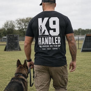 Puede incluir: Una camiseta negra de manga corta con texto blanco en la espalda que dice K9 HANDLER, WORKING DOG TEAM y TRAIN TRACK CONTROL, junto a un gráfico de una persona y un perro. Se combina con pantalones cargo de color caqui y una gorra negra.