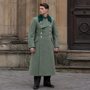 May include: A long, olive green coat with a double-breasted design and silver buttons. The coat features a dark green collar and is worn with dark pants and black shoes. The coat is a classic military style.