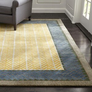 Può includere: Un tappeto rettangolare con un motivo a spina di pesce giallo e bianco al centro, incorniciato da un bordo blu con un design geometrico. Il tappeto ha un bordo dai toni neutri ed è posizionato su un pavimento in legno scuro.