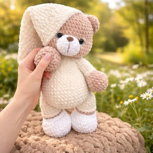 Może przedstawiać: Ręcznie robiony szydełkowy miś w kremowej piżamie i czapce. Miś ma brązowe akcenty na łapach, uszach i nosie. Miś ma około 25 cm wysokości i trzyma małą piłkę.