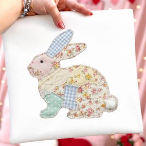 Może przedstawiać: Biała kwadratowa poduszka z patchworkowym wzorem kr&oacute;lika. Kr&oacute;lik wykonany jest z r&oacute;żnych tkanin, w tym nadruk&oacute;w kwiatowych, kraty vichy i koronki, w pastelowych kolorach. Kr&oacute;lik ma r&oacute;żową twarz i uszy oraz puszysty biały ogon.