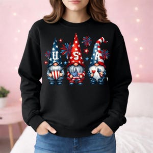 May include: Black sweatshirt featuring three patriotic gnomes with the letters "USA". The gnomes are dressed in red, white, and blue, holding flags and hearts, with fireworks in the background. A festive design for Independence Day.