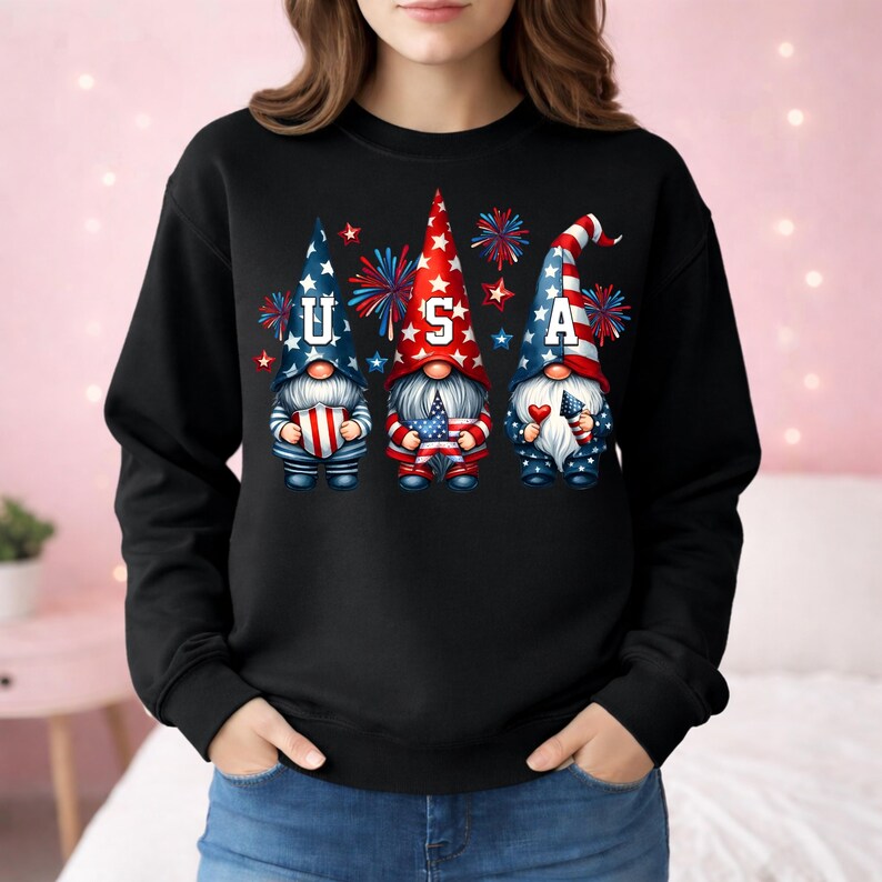 May include: Black sweatshirt featuring three patriotic gnomes with the letters "USA". The gnomes are dressed in red, white, and blue, holding flags and hearts, with fireworks in the background. A festive design for Independence Day.