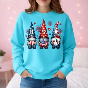 May include: A light blue sweatshirt featuring three gnomes with patriotic designs. Each gnome wears a tall hat with the letters "U", "S", and "A". The gnomes hold flags and fireworks are in the background. The sweatshirt is a solid light blue color.